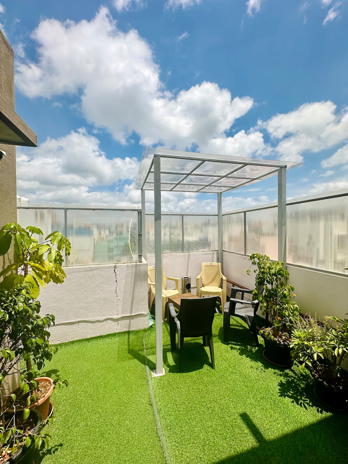 A private outdoor terrace garden is shown, featuring a clear canopy overhead and a green artificial lawn. A table and several chairs are surrounded by potted plants, creating a serene space for relaxation and enjoying the city views.