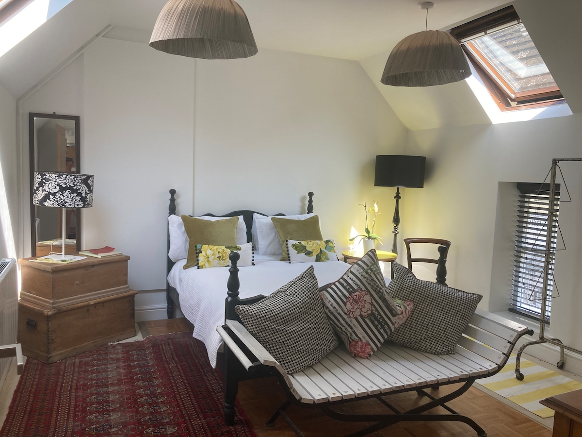 A bright double bedroom features a spacious bed adorned with decorative pillows. Natural light filters through Velux windows, illuminating the whitewashed walls and wooden floor. A wooden trunk and a vintage chair add character, while a striped lounge chair offers a cozy spot for relaxation.