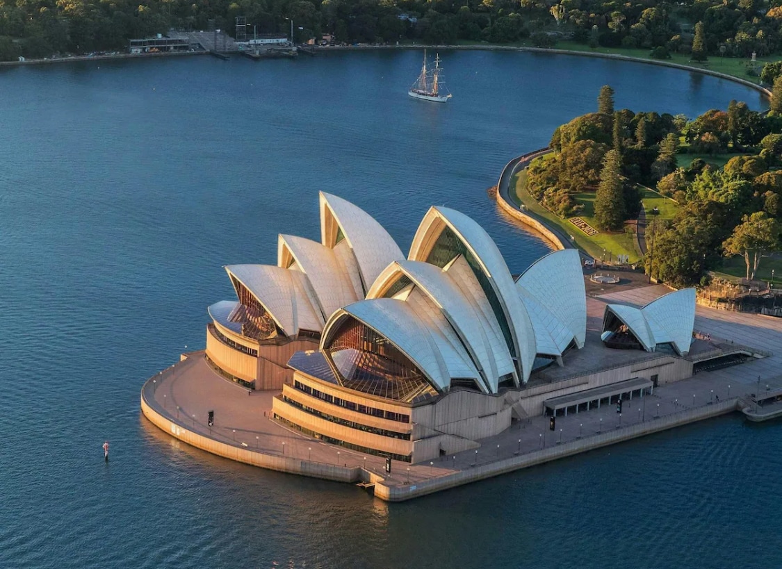 The Sydney Opera House, characterized by its distinctive sail-like structures, is featured prominently against the backdrop of Sydney Harbour. The surrounding water reflects the building's shapes, while greenery and pathways can be seen along the shoreline.