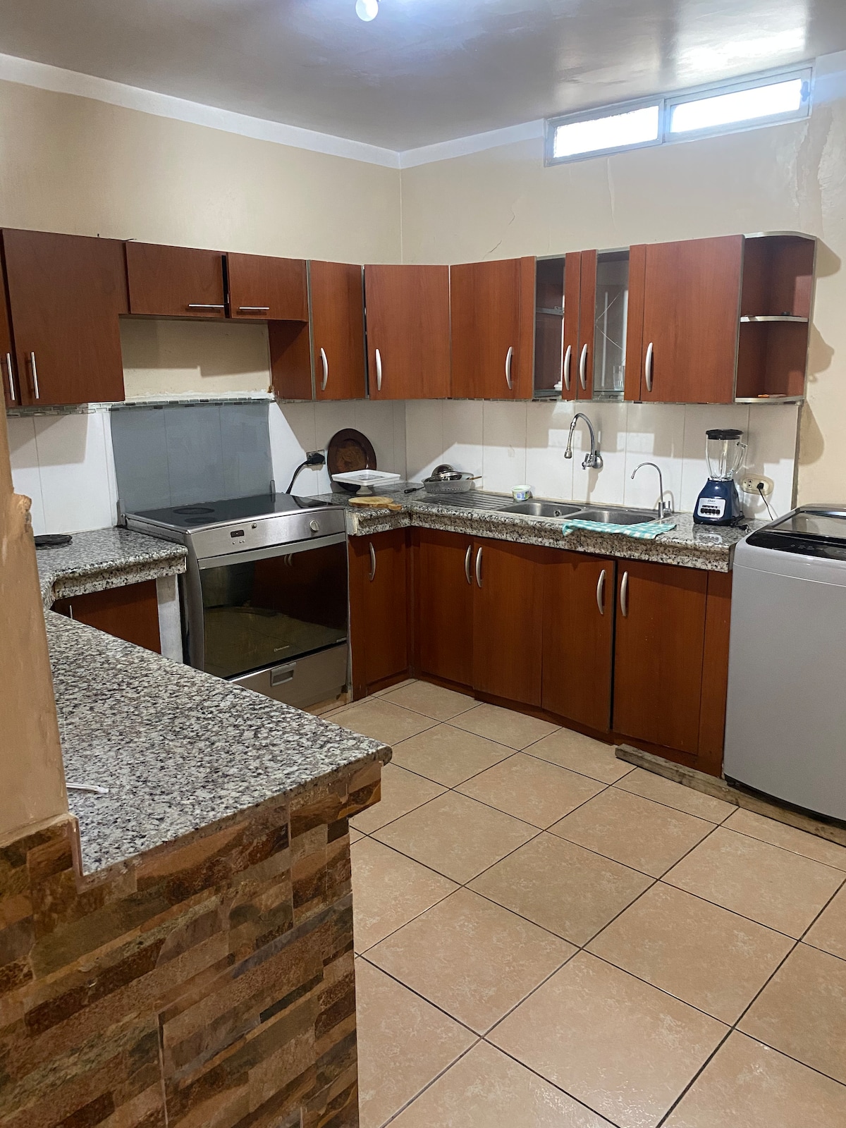 The kitchen features wooden cabinetry with a granite countertop. Equipped appliances include a stainless steel dishwasher and a blender, along with a sink and a washing machine. Natural light enters through a small window, enhancing the functional workspace.