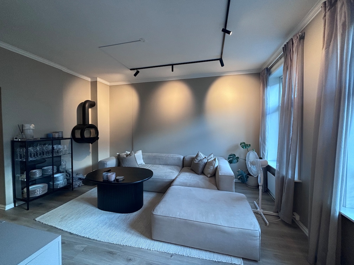 A spacious living area is presented, featuring a light-colored sectional sofa arranged around a circular black coffee table. Soft natural light filters through large windows, illuminating the room. A plant and a fan are positioned nearby, adding to the room's functionality.