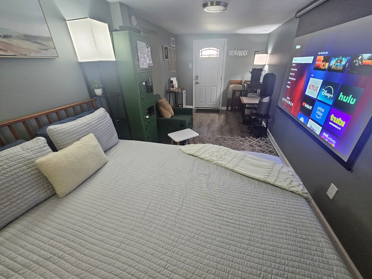A cozy studio workspace is displayed, featuring a spacious queen bed with soft linens. A wall-mounted screen shows various streaming services. The entrance door is visible, complemented by a small desk and chair in the corner. A light gray color palette is used throughout.