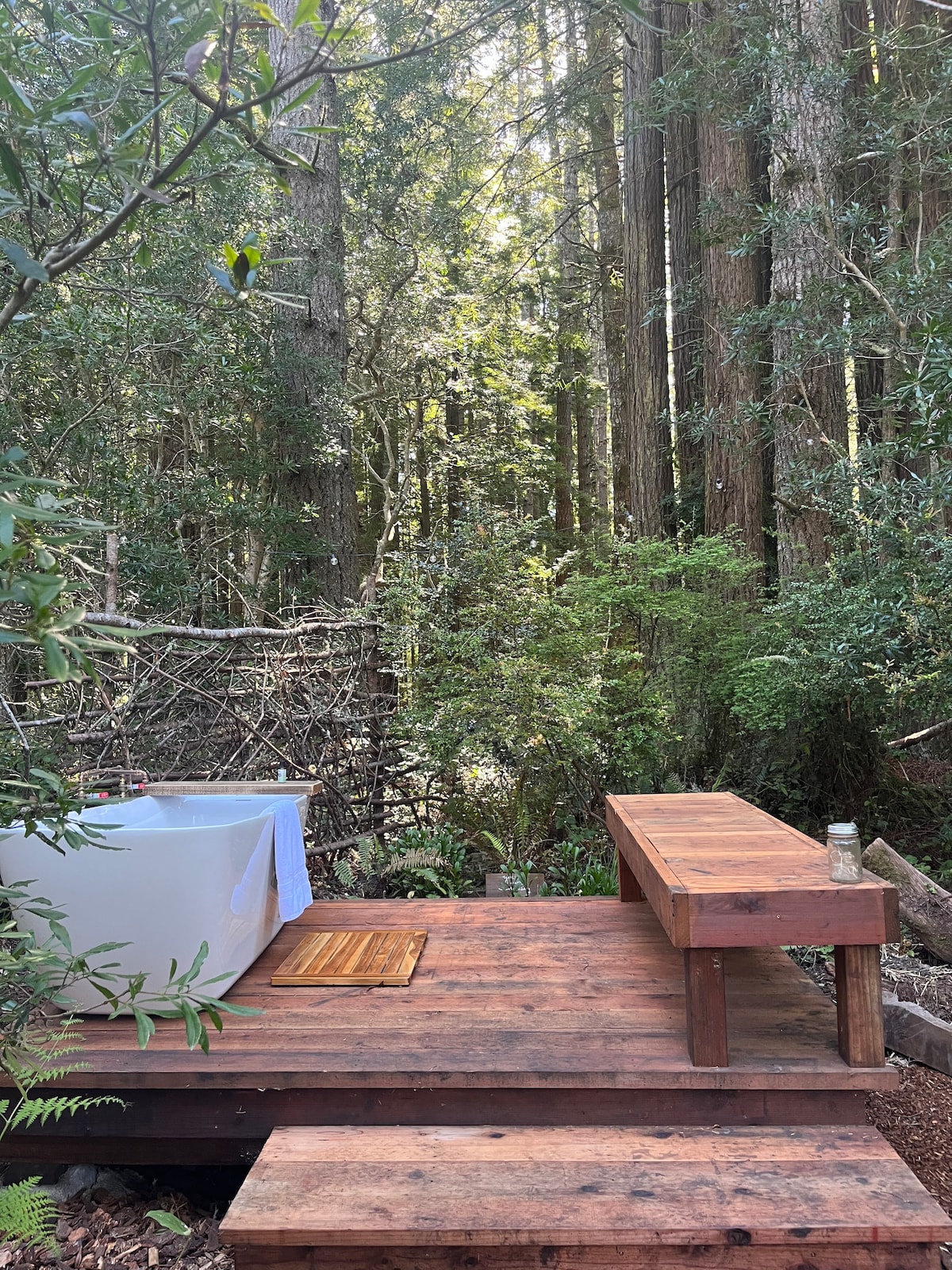 An outdoor bathing area is nestled among towering trees, featuring a freestanding tub on a wooden deck. A simple wooden bench is positioned nearby. Lush greenery surrounds the space, creating a serene and private atmosphere.