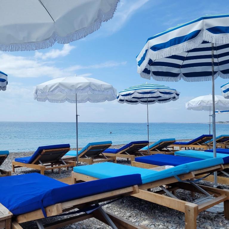 A collection of beach loungers is arranged under large, blue and white striped umbrellas along a pebbly shoreline. The calm sea stretches into the distance, meeting a cloudy sky, creating a serene coastal environment.