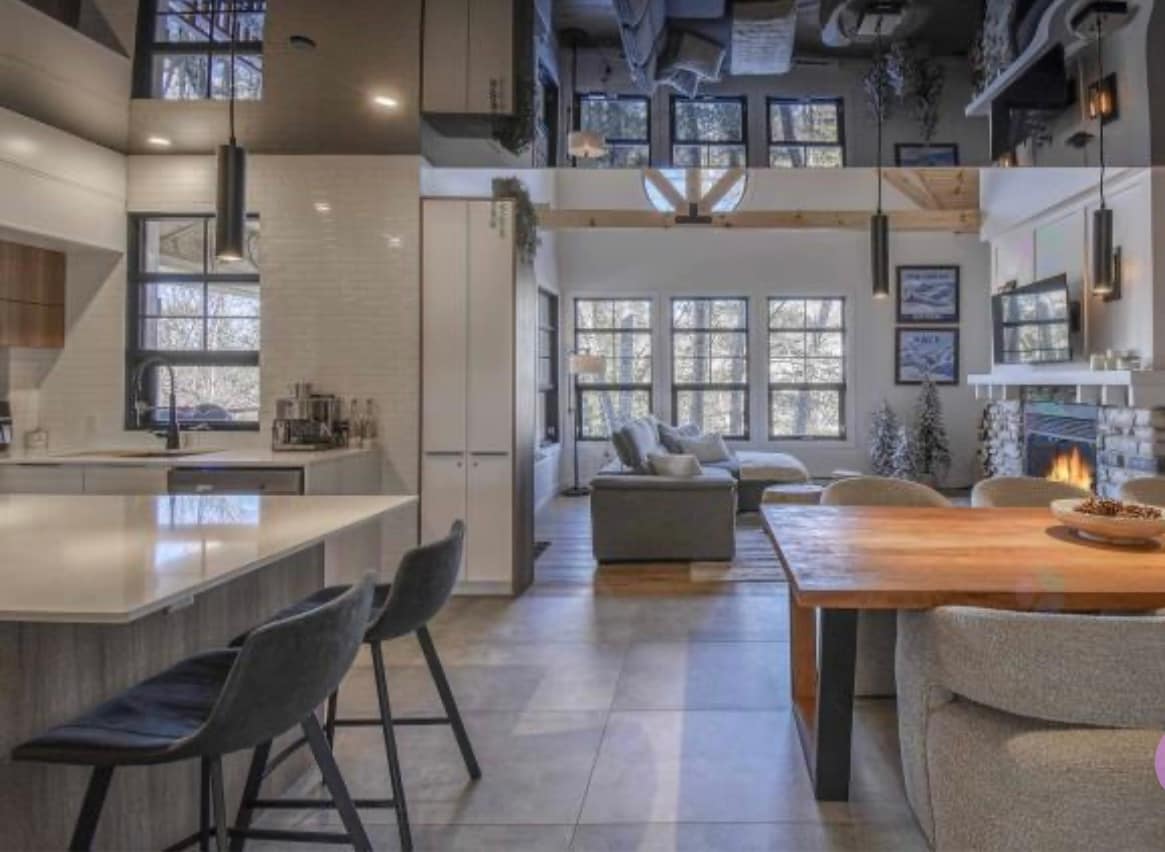 An open-concept living space is displayed, featuring a modern kitchen with an island and a dining area with a wooden table. The seating area includes a spacious couch and is illuminated by natural light streaming through large windows, reflecting a cozy ambiance complemented by a fireplace.