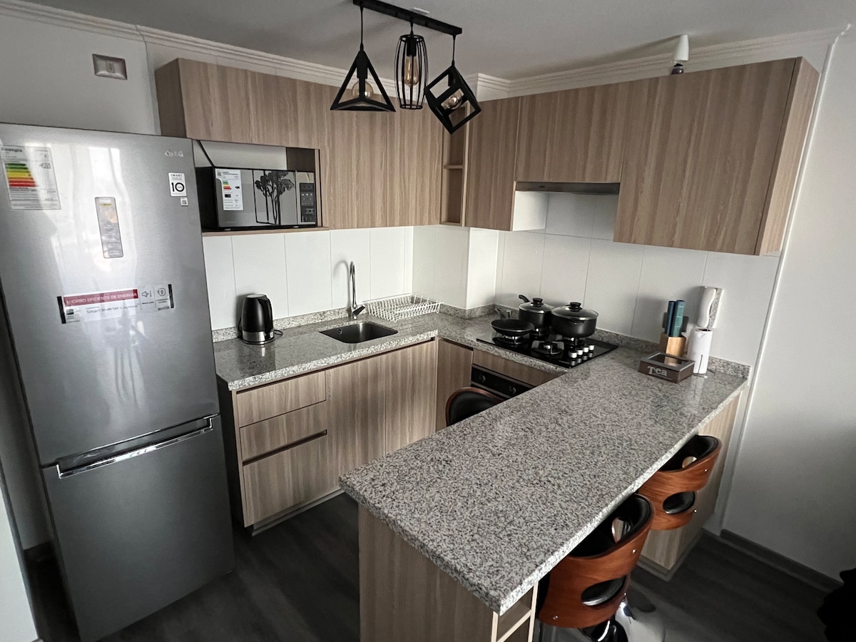 The kitchen features a modern design with wooden cabinetry and a granite countertop. Equipped with a stainless steel refrigerator, microwave, and a gas stove, the space offers a breakfast bar with three wooden stools. Natural light brightens the room through a nearby window.
