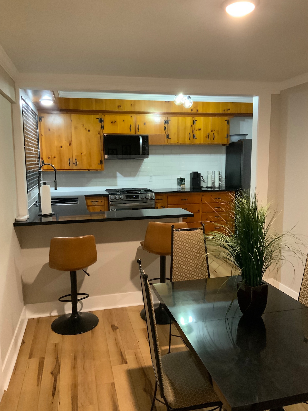 The kitchen features wooden cabinetry and modern appliances, including a gas stove and microwave. A small dining area is visible, equipped with a black table and four chairs. Two bar stools are positioned at the kitchen countertop, accented by a potted plant.