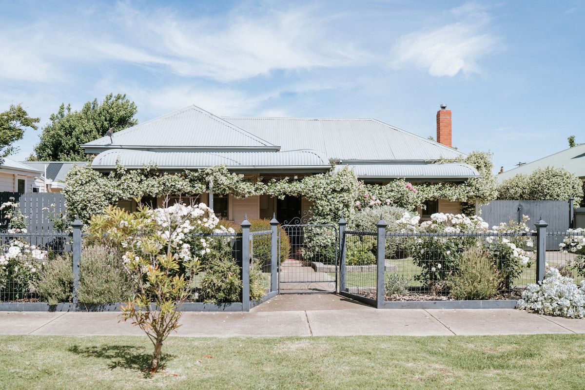 A charming residence is framed by a well-maintained garden, featuring lush greenery and flowering shrubs. The covered porch is adorned with climbing vines, creating an inviting entrance. The house's classic design is highlighted by a metal roof and warm wooden accents.