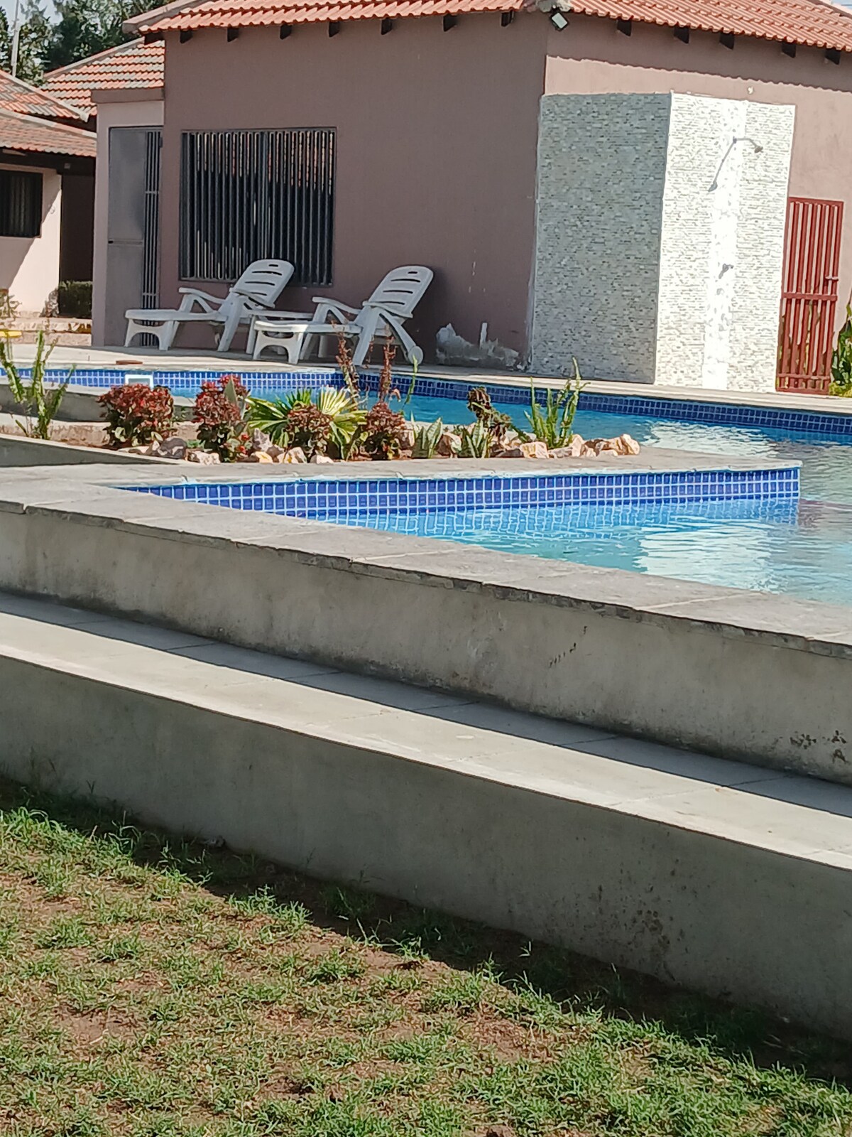 A serene pool area is framed by landscaped beds featuring a variety of plants. Light-colored lounge chairs are positioned nearby, while a simple structure with a textured wall stands in the background, creating a restful environment for guests.