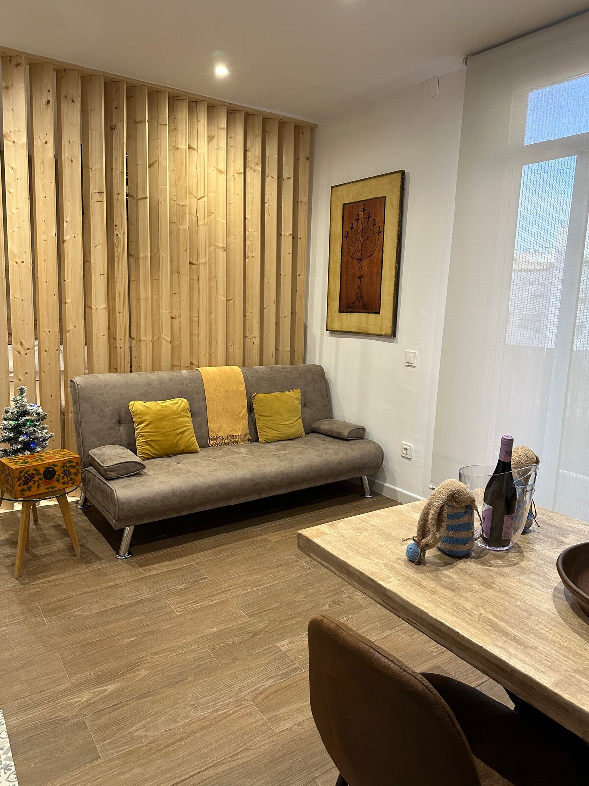 A modern living area features a comfortable sofa bed accented with yellow pillows, positioned beside a wooden wall with vertical slats. A dining table is present, adorned with a decorative bowl and a small Christmas tree, adding a touch of warmth to the space.