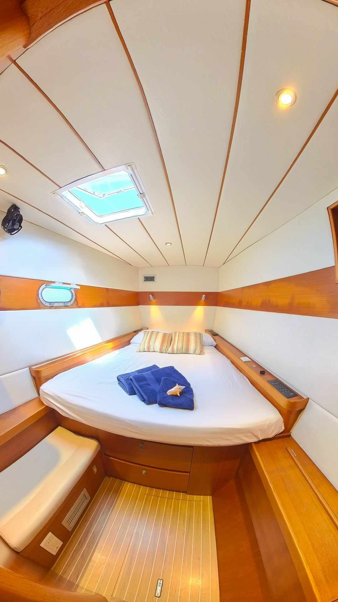 A cozy cabin features a spacious circular bed with striped bedding and a soft blue towel folded on top. Natural light enters through a skylight above, complementing the warm wooden accents and light-colored walls. A small seating area is visible to the side.