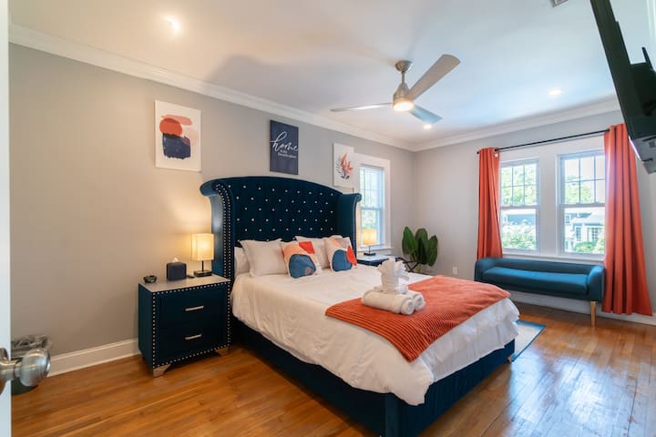 This high-end bedroom features a magnificent navy tufted velvet bed, matching designer nightstands, a large flat screen TV, and a chic window bench. Warm terracotta accents and rich hardwood floors complete the elite vibe.