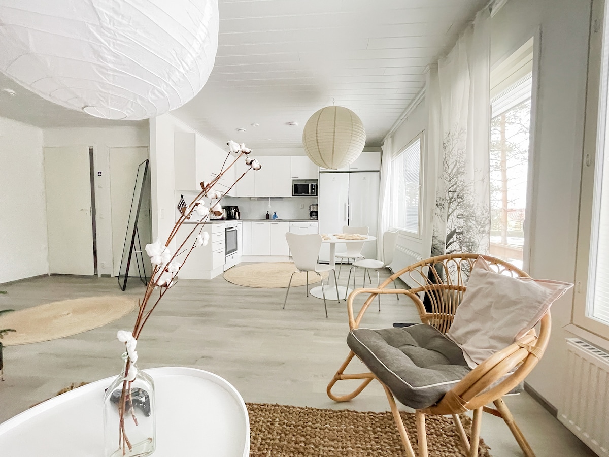 A modern, light-filled living area features a stylish kitchen and dining space. Natural elements are showcased, including a decorative branch and a soft area rug. Large windows allow for ample daylight, while a cozy armchair provides a relaxing seating option.