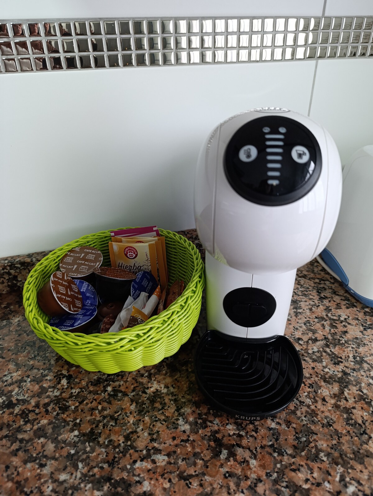 A compact coffee machine is positioned next to a woven, green basket filled with various coffee pods, creamers, and sweeteners. The countertop features a smooth granite surface, enhancing the kitchen's functionality.