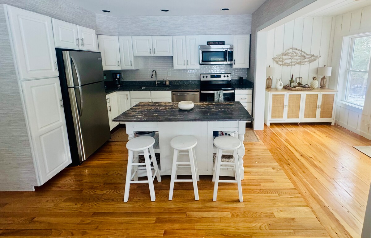 A modern kitchen features white cabinetry, sleek stainless steel appliances, and a central island with a dark countertop. Three white stools are positioned at the island. Warm wooden floors extend throughout the space, enhancing the light and airy atmosphere.