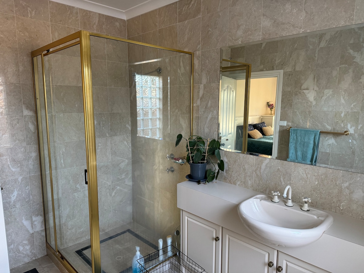A bright bathroom features a glass shower enclosure with gold trim, complemented by marble-effect walls. A large mirror reflects the space. A sleek vanity with a round sink and two faucets is positioned beneath the mirror, alongside a potted plant adding a touch of greenery.
