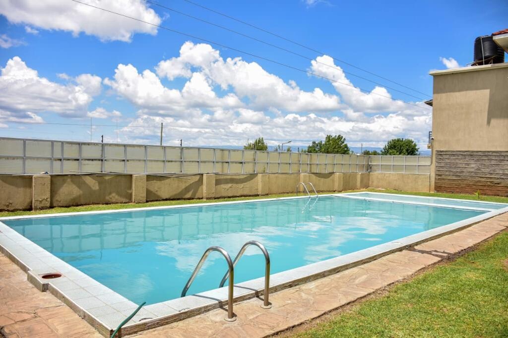 A sunny outdoor pool is featured, surrounded by a neat stone deck and grassy areas. The clear blue water reflects the sky and clouds above, while a low wall provides some privacy from the surrounding environment.