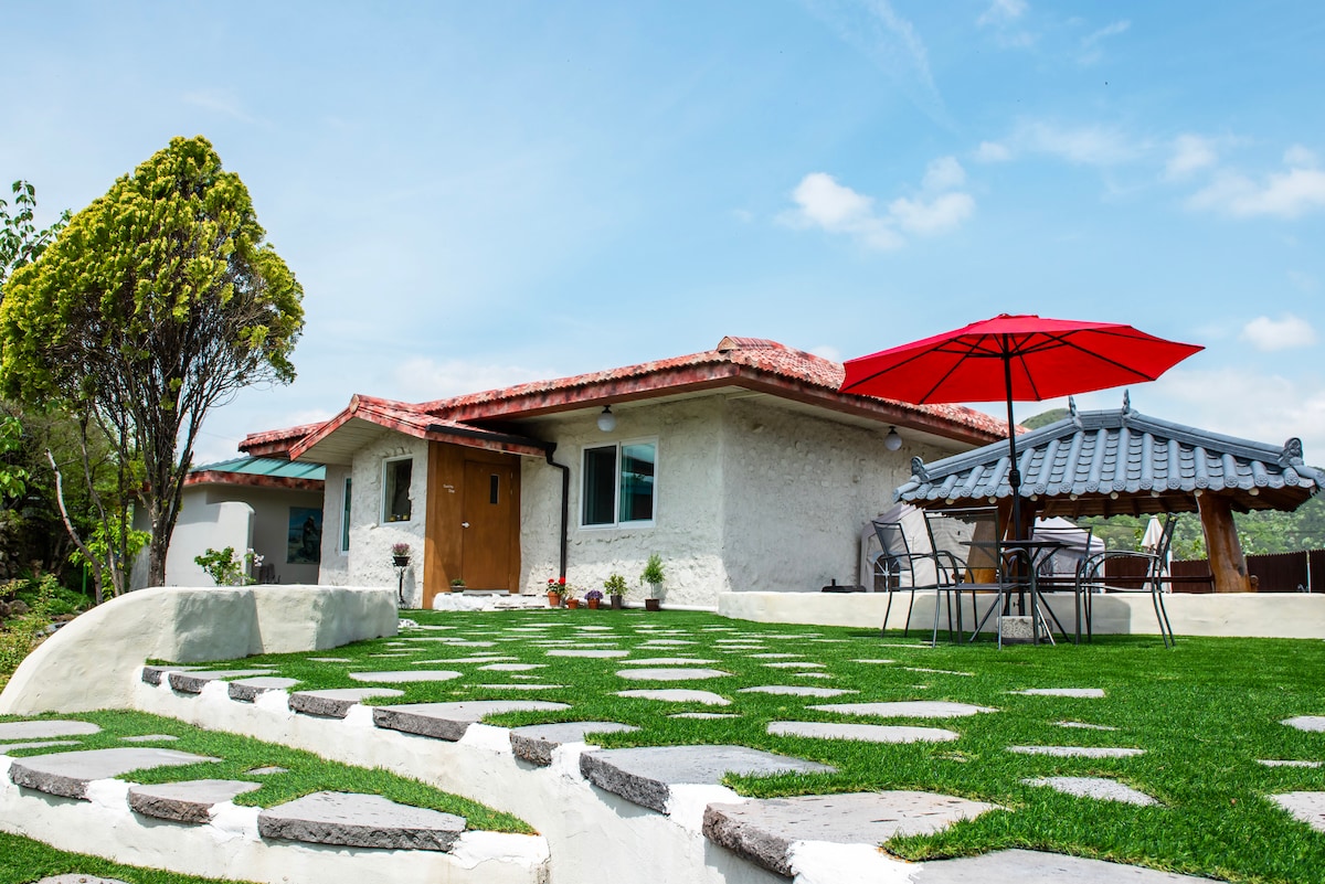 A charming building with Mediterranean-inspired architecture is surrounded by lush greenery. A spacious patio area features a red umbrella providing shade over a table and chairs, while stone steps lead up to the entrance of the home.