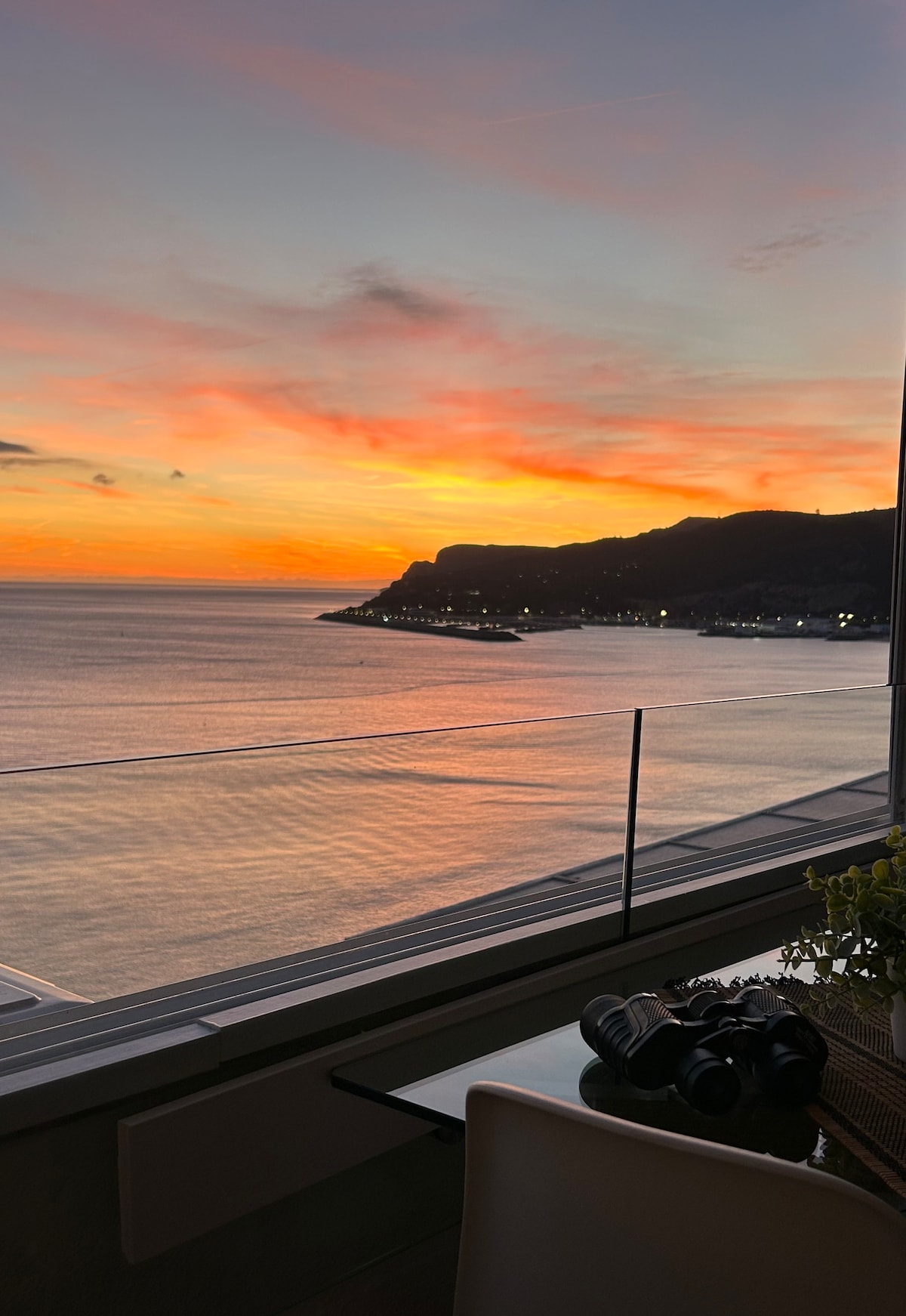 A breathtaking sunset is showcased over the ocean, casting warm hues of orange and pink across the water. The rugged coastline is visible in the background, illuminated by distant lights. A clear glass railing frames the view, enhancing the connection with the serene landscape.