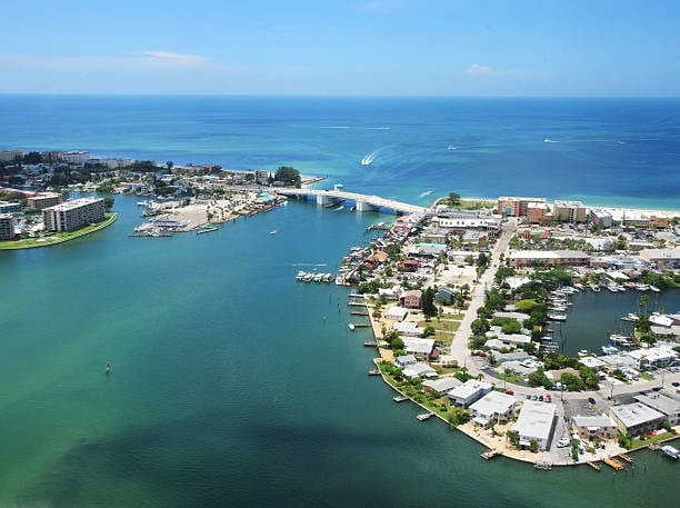 An aerial view captures the vibrant coastal area featuring the shimmering blue waters of the Gulf of Mexico. A bridge connects the marina to the nearby shoreline, where boats are moored. Lush greenery and residential buildings are visible along the waterfront, creating a scenic coastal landscape.