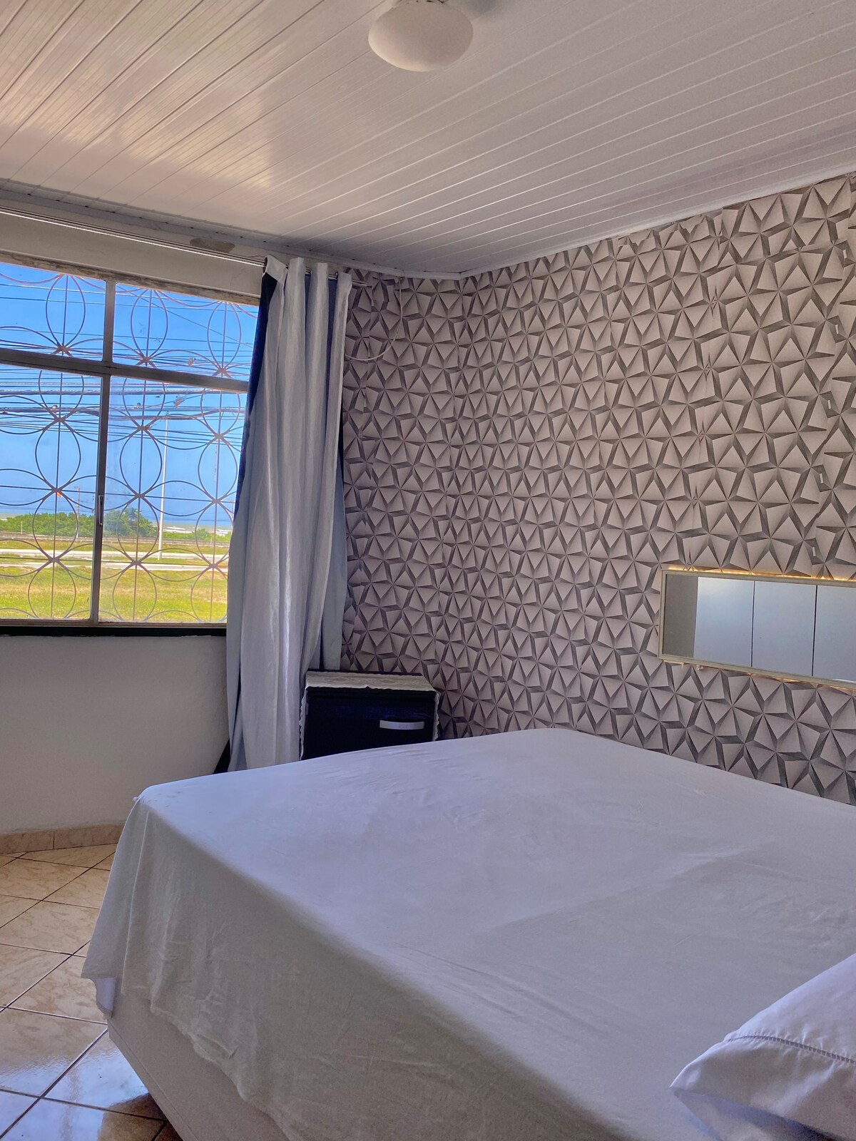 A bright bedroom features geometric patterned wallpaper and a spacious bed covered with a white sheet. Soft natural light enters through a large window, offering a view of the surrounding area. A mirrored wall piece reflects the room's design elements.