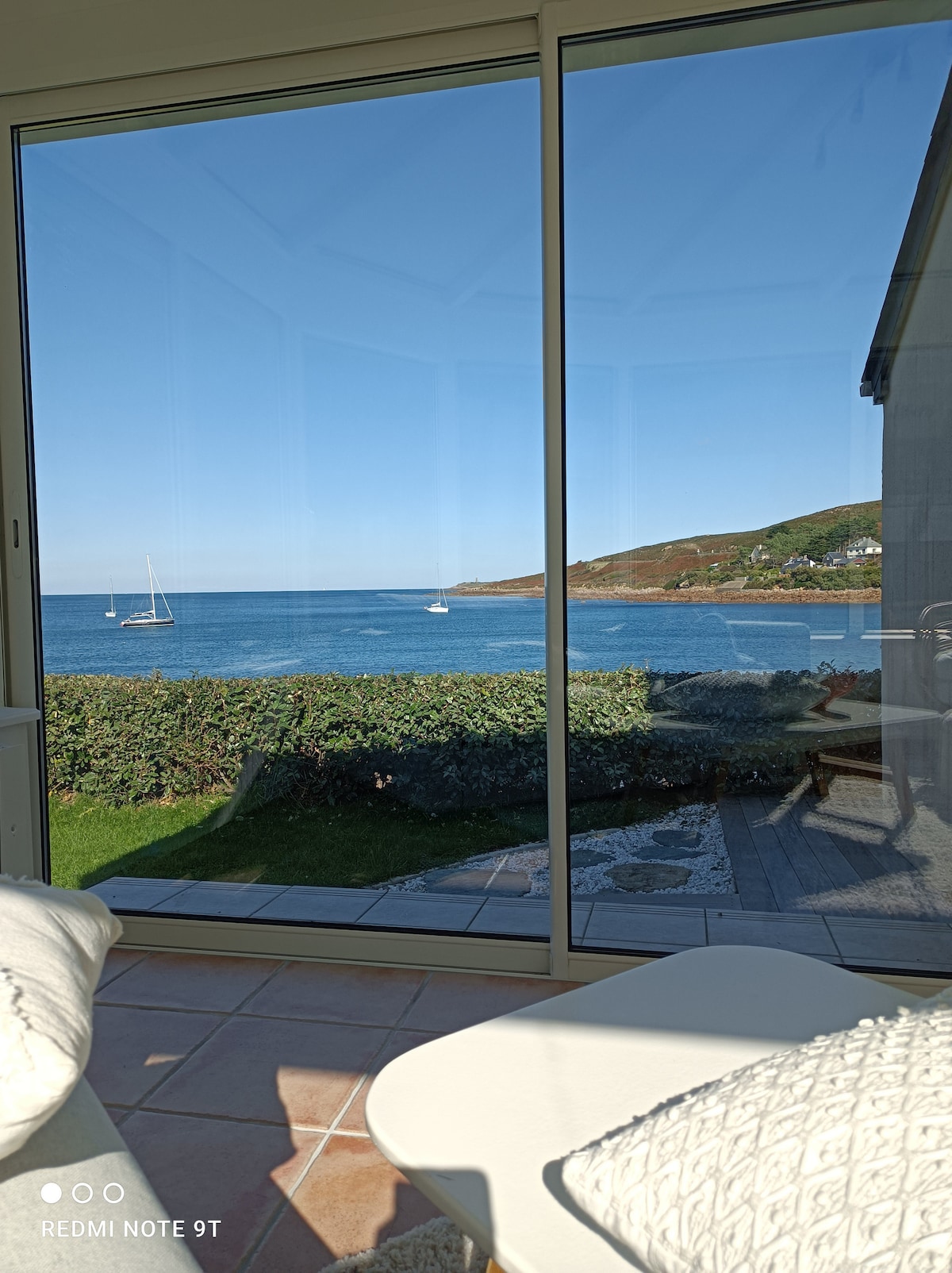 A large glass window frames a serene view of the coastline, with sailboats visible on the calm water. Lush green vegetation and a patio area are seen in the foreground, while gentle hills rise in the background under a clear blue sky.
