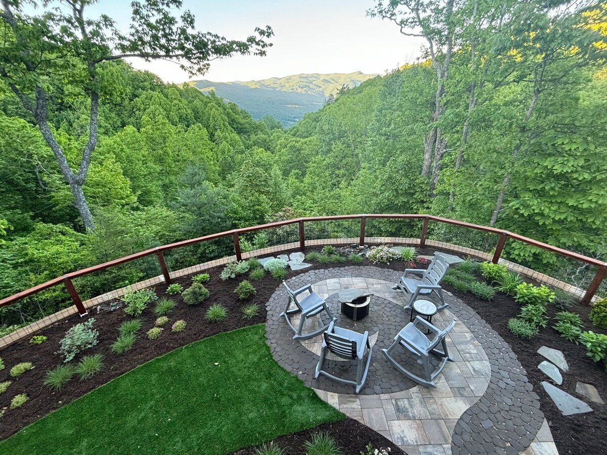 An outdoor terrace area is featured, surrounded by lush green trees and mountains in the distance. A circular stone patio holds a fire pit, accompanied by multiple chairs arranged for seating. Various plants and landscaping elements add to the serene setting.