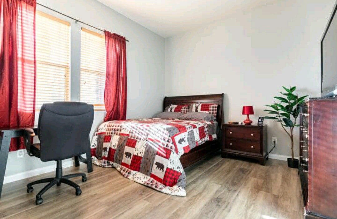 A spacious bedroom features a queen bed adorned with a patterned quilt, complemented by red curtains framing two windows. A sturdy desk with an ergonomic chair is positioned nearby, alongside a dresser and a 43" TV, enhancing the room's functionality.