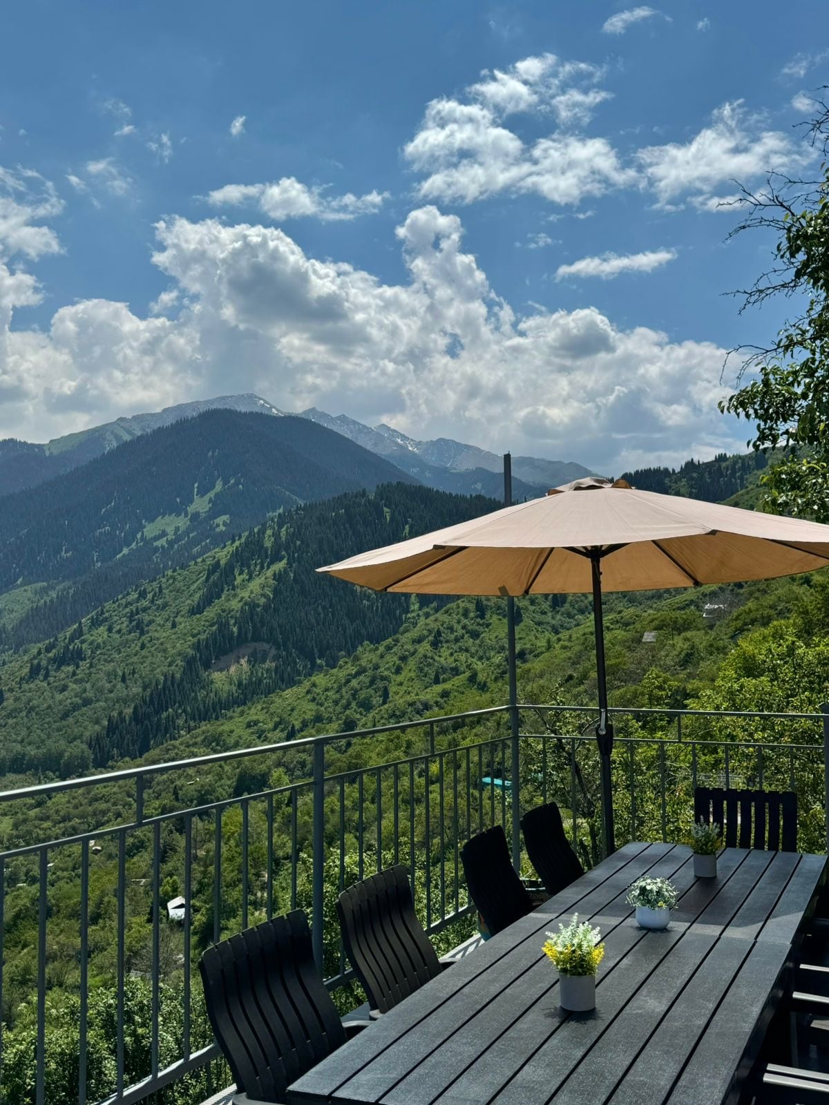 An outdoor dining area is situated on a terrace, featuring a long table surrounded by chairs. A large umbrella provides shade while beautiful mountain views are visible in the background, showcasing rolling hills and lush greenery under a partly cloudy sky.