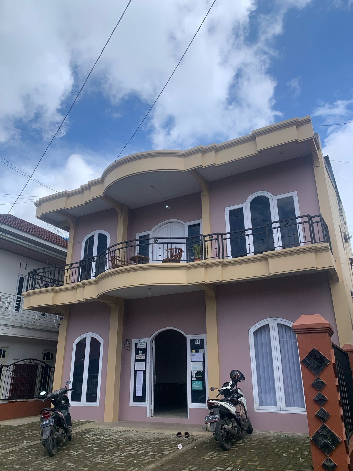 A two-story building is shown with a light pink exterior and white trim. Balconies are visible on the upper level, and large arched windows punctuate the facade. A motorcycle is parked in front, while the sky above is partly cloudy.
