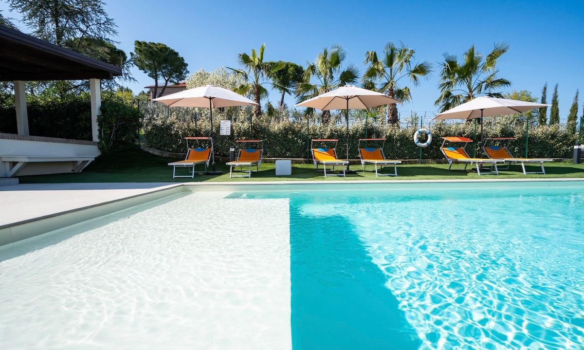 A sparkling swimming pool features shallow steps leading into the water. Loungers with vibrant orange upholstery are arranged around the pool, shaded by large umbrellas. Lush palm trees and greenery create a serene outdoor setting in the background.