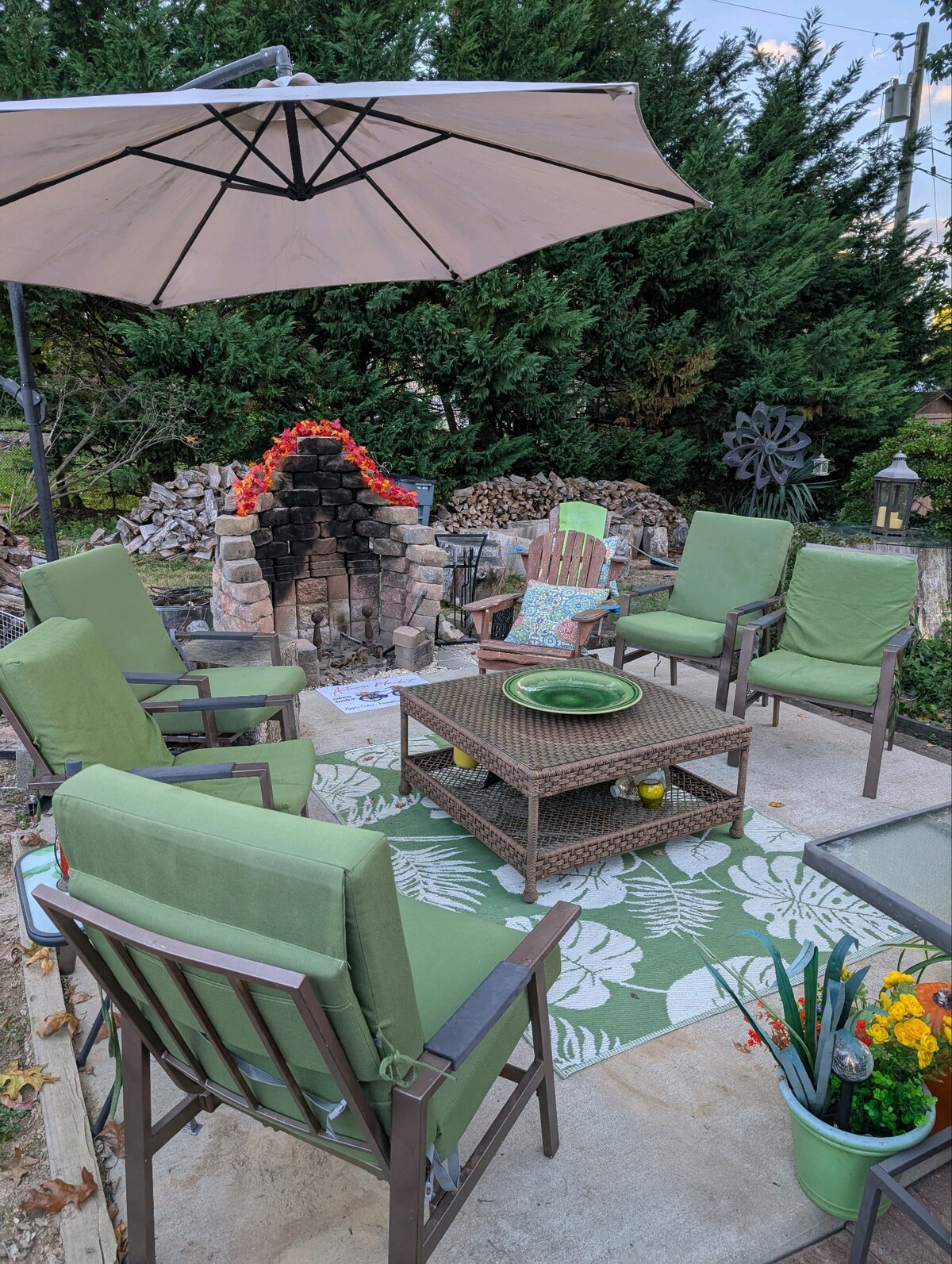 An outdoor seating area is presented with six green chairs arranged around a central coffee table. A decorative area rug with leaf patterns is placed on the concrete surface. An umbrella provides shade, and a stone fire pit adds a cozy element in the background.