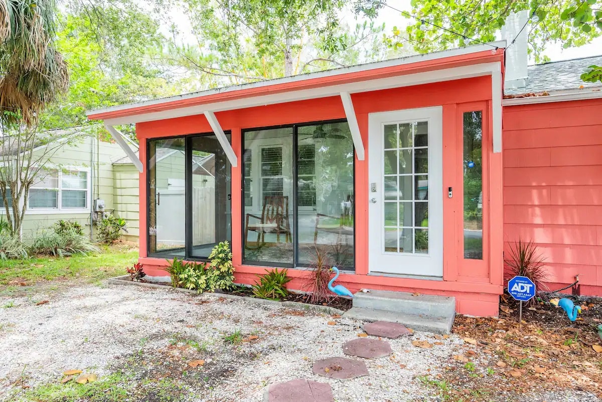 The cottage exterior features a coral-colored facade with large glass sliding doors that provide natural light. A welcoming entryway leads to a covered porch area with a rocking chair, surrounded by lush greenery and well-maintained landscaping.