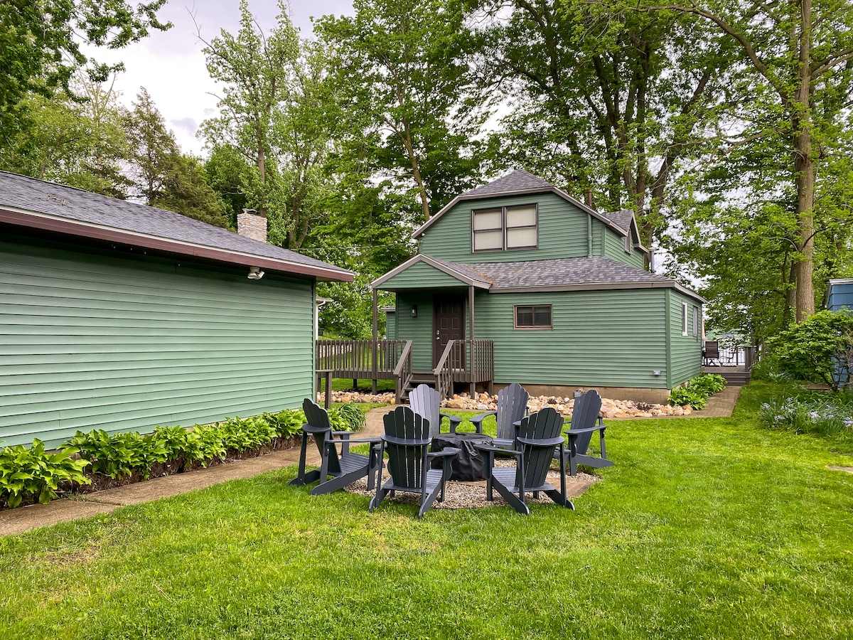 A charming green cabin is depicted, nestled among lush trees. A fire pit area with adirondack chairs is arranged in a circular layout, surrounded by a well-kept lawn. The welcoming entrance features a small set of steps leading to the door, enhancing the outdoor space.