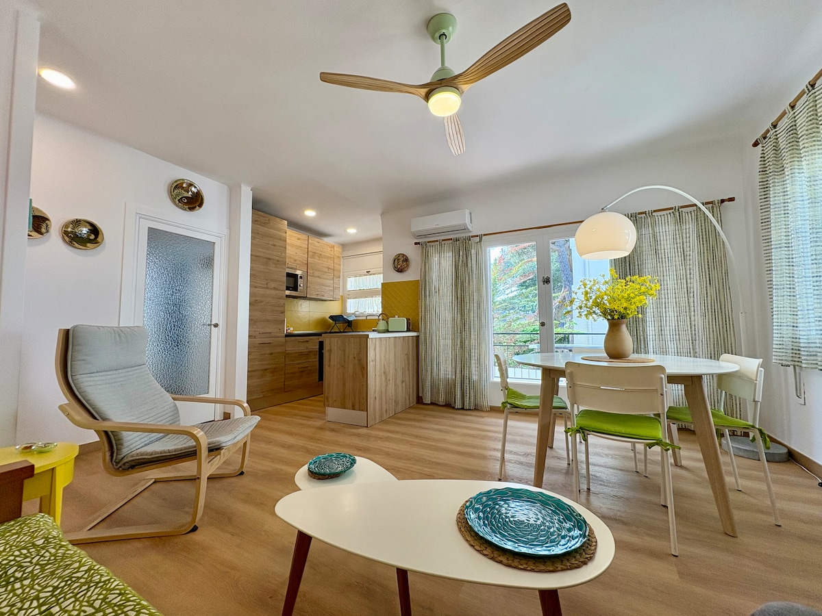 An open-plan living area presents a modern decor. A dining table with colorful chairs is set beside a well-equipped kitchen. A light fixture hangs gracefully over the table. Natural light filters through the glass doors, leading to a balcony with garden views.