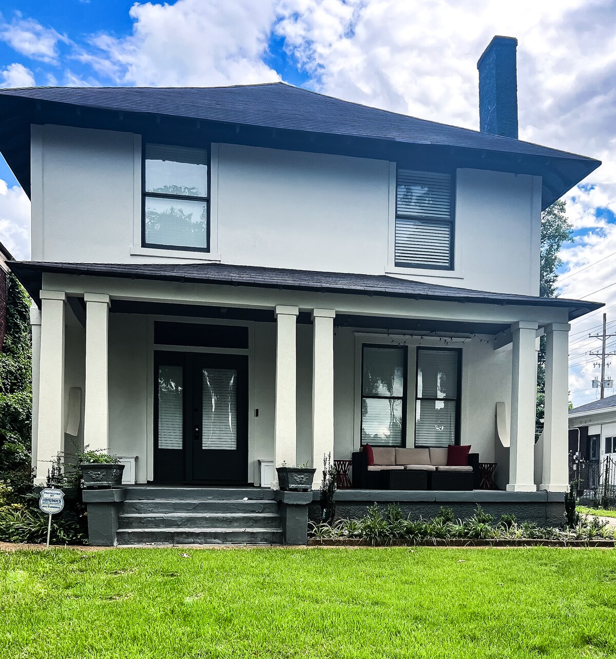 A two-story home with a classic design is featured, showcasing a welcoming front porch supported by tall columns. A neatly manicured lawn surrounds the house, and comfortable outdoor seating is arranged on the porch. Large windows invite natural light into the space.
