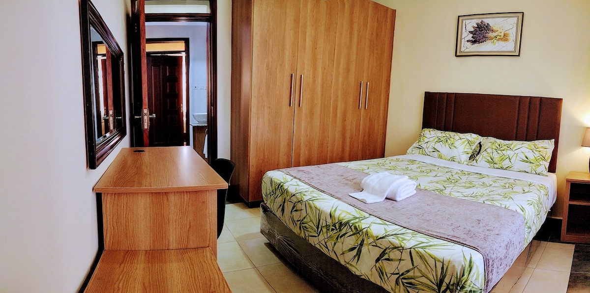 A spacious bedroom is featured, with a large bed adorned in a floral-patterned bedding set. A wooden desk is positioned adjacent, and a mirror is mounted above it. Built-in wardrobes complement the design, providing ample storage space. Soft lighting creates a calm ambiance.