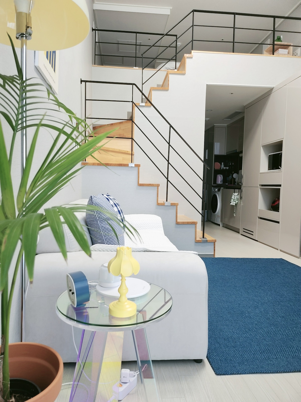 A modern living area is displayed with a light-colored sofa facing a blue area rug. A small glass table is positioned nearby, featuring a yellow lamp and decorative items. A wooden staircase leads to an upper level, with sleek railings adding to the contemporary design.