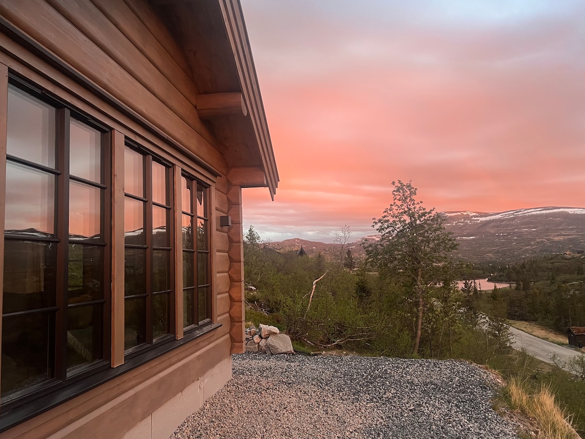 A corner of the timber cabin is captured at sunset, reflecting warm hues in the large windows. The surrounding landscape features greenery and mountains, providing an expansive view of the natural scenery, while a gravel area leads up to the cabin's porch.