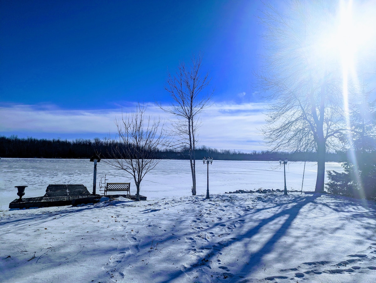 A tranquil winter scene showcases a frozen river, with snow-covered ground and clear blue skies. Leafless trees stand alongside a wooden bench and a stone platform that leads to the water's edge. Shadows are cast by the trees, creating a serene atmosphere.