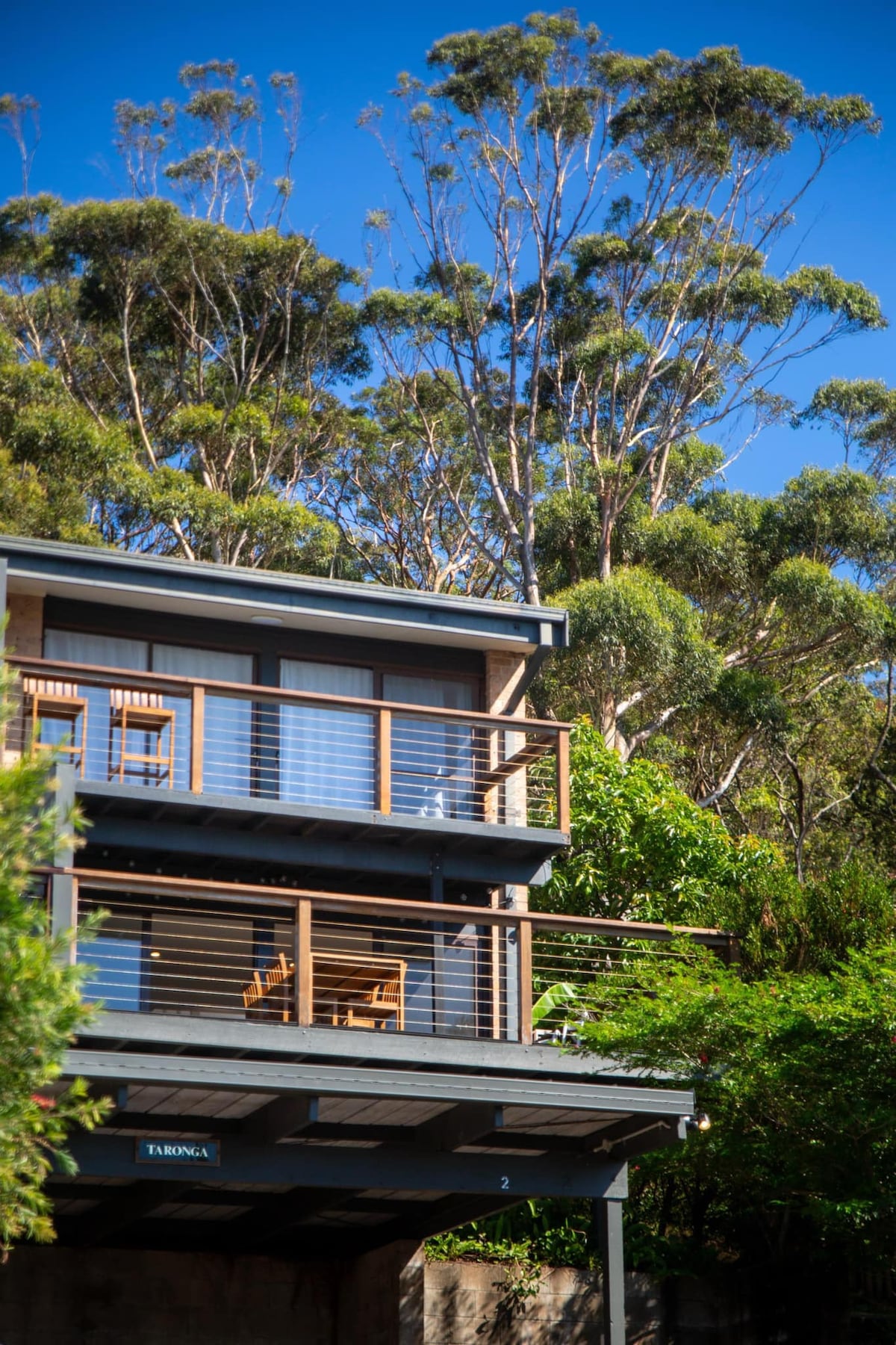 A two-story duplex is seen set amongst lush greenery, featuring a balcony with two wooden chairs. The exterior showcases large windows that invite natural light, while tall trees in the background enhance the tranquil ambiance of the surrounding area.