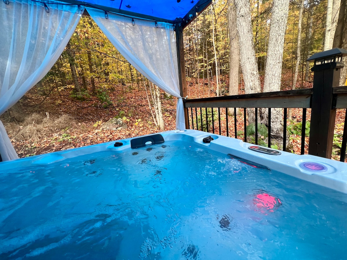 A hot tub is set on a wooden deck, surrounded by sheer curtains. The vibrant blue water reflects the natural light, with trees adorned in autumn foliage visible in the background, creating a peaceful outdoor retreat.