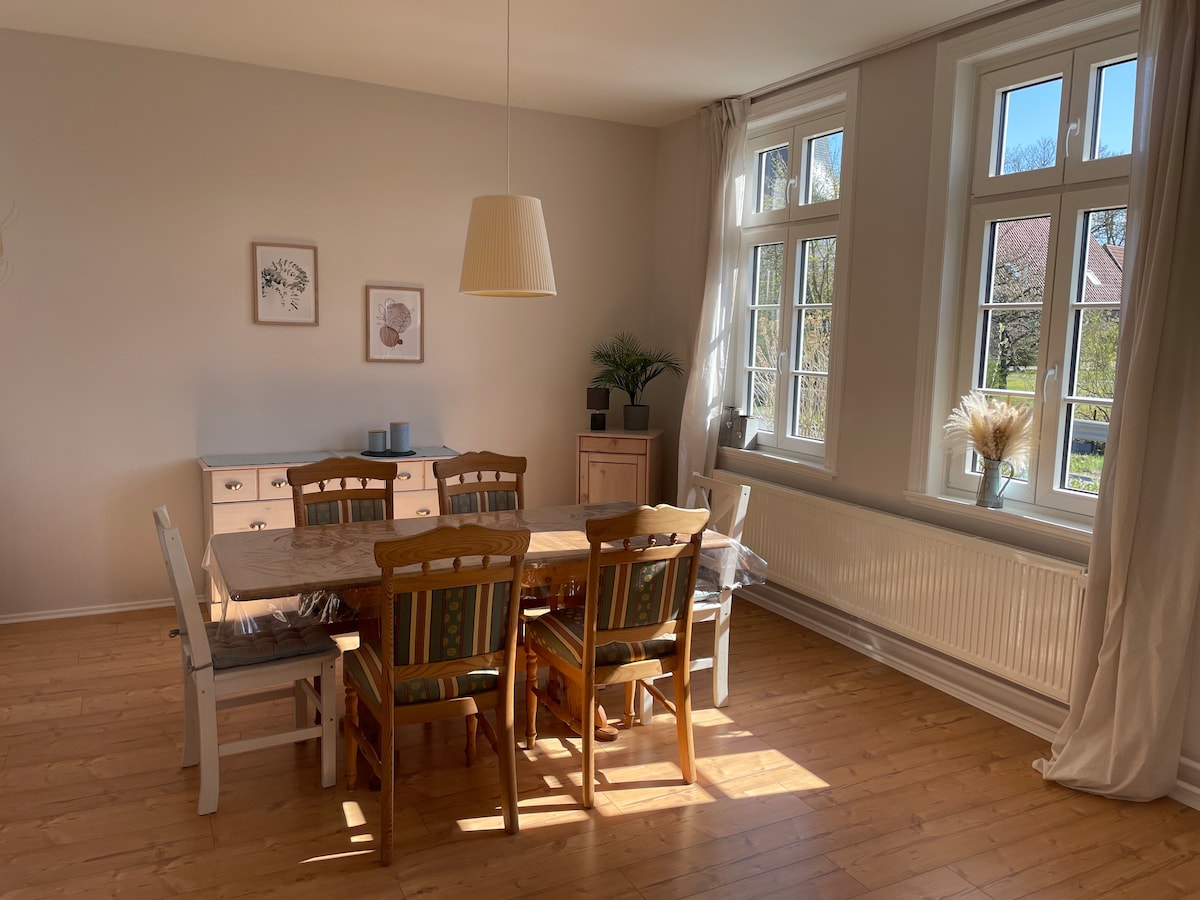 A dining area is featured, showcasing a wooden table surrounded by six chairs. Large windows allow natural light to fill the space, highlighting the warm wooden flooring. A sideboard is present, complemented by minimal decor and potted greenery.