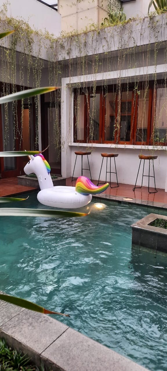 A unicorn-shaped inflatable floats in the turquoise water of a private swimming pool. Bar stools are positioned along a counter in the background, complemented by large windows that allow natural light to fill the space.