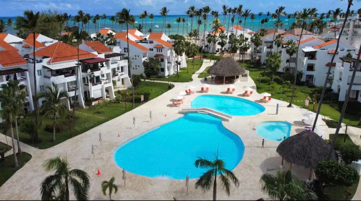 The image showcases a spacious outdoor area with a large swimming pool, surrounded by palm trees and lush greenery. Relaxing lounge chairs are positioned along the poolside. In the background, the ocean is visible, framed by charming white buildings with red roofs.