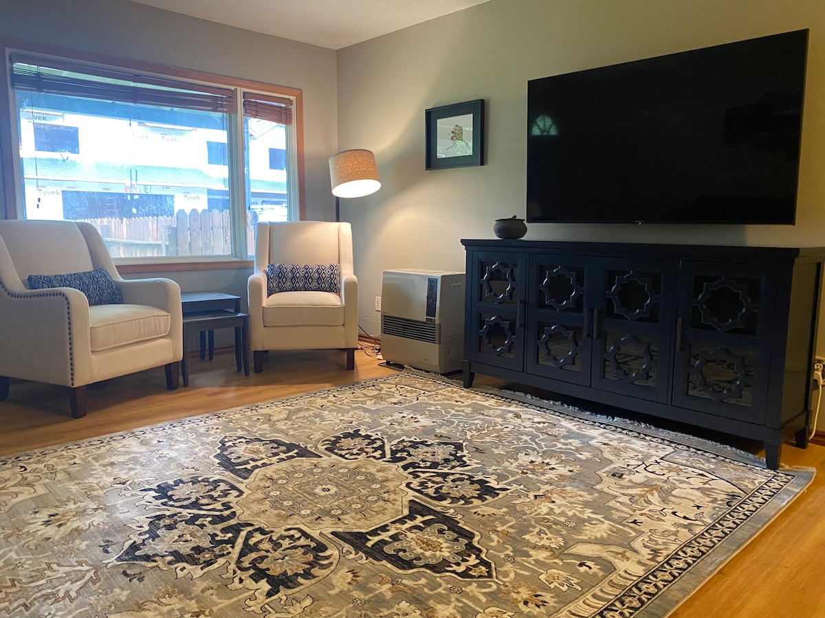 A cozy living area features two armchairs positioned beside a decorative lamp. A large television is mounted on the wall across from a dark wood console. A patterned area rug, with neutral tones and intricate designs, enhances the warm ambiance of the space.