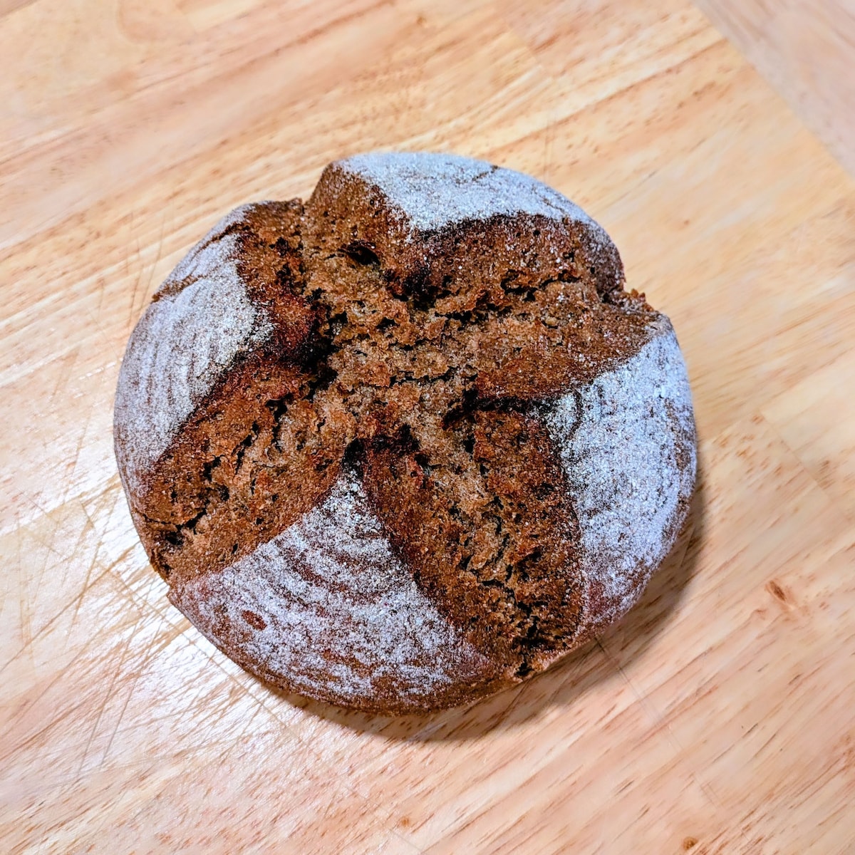 A round loaf of sourdough bread is displayed on a wooden surface. The crust is dark brown with a rustic texture, featuring a star-like pattern on the top. A light dusting of flour covers parts of the crust, highlighting its artisanal appearance.