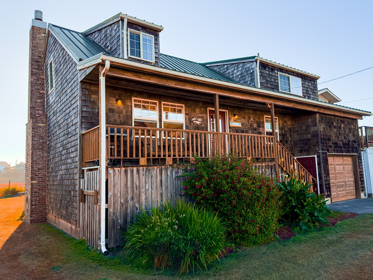A charming two-story river home features a wooden façade with a sloped metal roof. A spacious front porch, bordered by flowering plants, provides seating opportunities. Multiple windows allow natural light to fill the interior, and the home is set against a serene early morning backdrop.