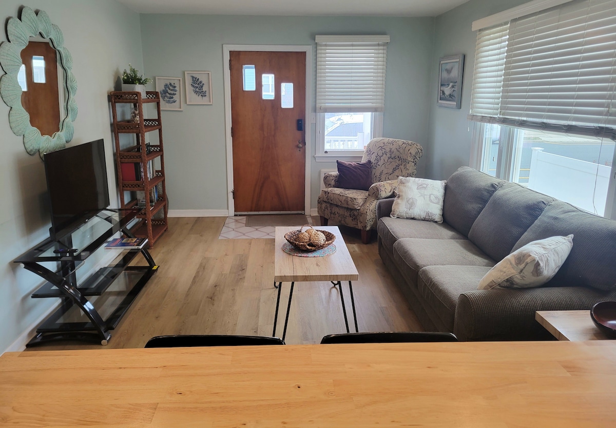 A comfortable living area is showcased with a gray couch and an armchair adorned with subtle patterns. A wooden coffee table sits in the center, complemented by a decorative bowl. A flat-screen TV is positioned on a stand against a light-colored wall, with natural light filtering in through the window.