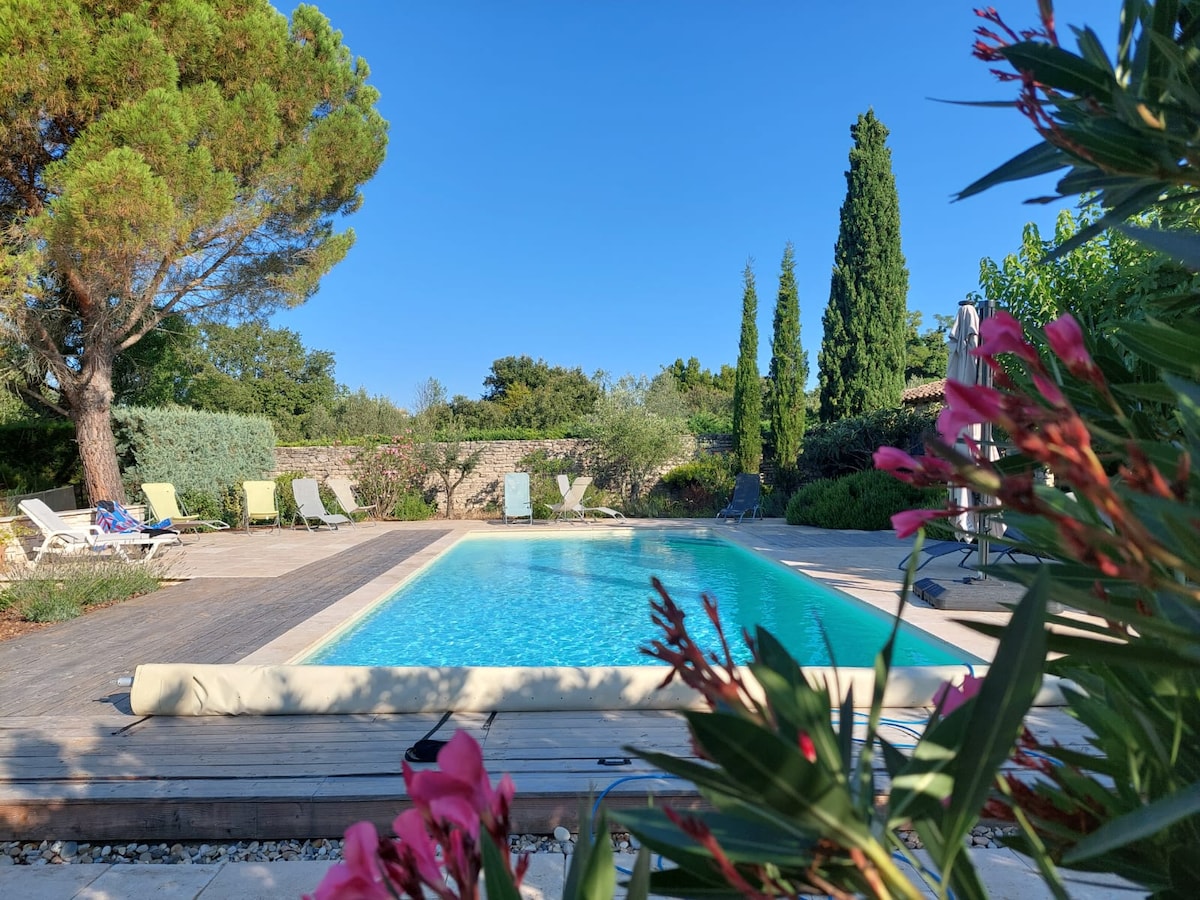 A refreshing outdoor pool is framed by a wooden deck and surrounded by lush greenery. Several lounge chairs are positioned around the pool area, offering space for relaxation. Vibrant pink flowers in the foreground add a touch of color to the serene landscape.